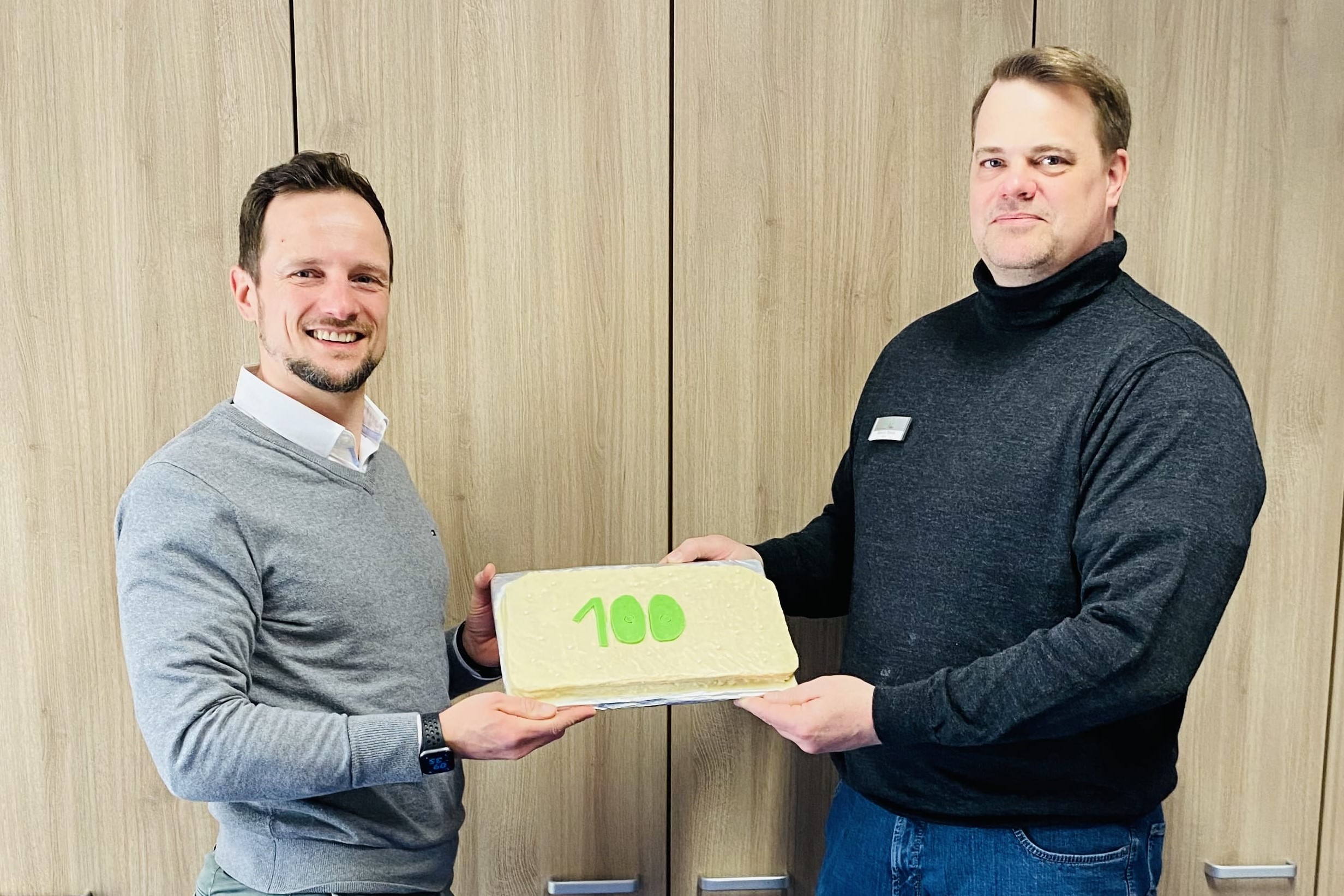 Zwei Männer stehen vor einem Holzschrank und halten gemeinsam eine rechteckige Torte mit grüner Aufschrift „100“. Beide lächeln in die Kamera – die Szene wirkt feierlich und deutet auf ein Jubiläum oder einen besonderen Anlass hin.